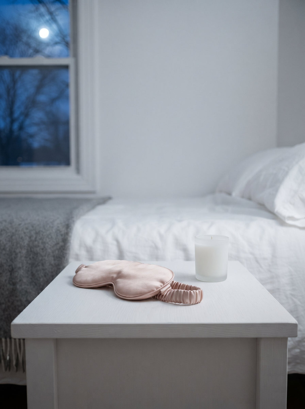A blush satin eye mask and a white candle on a bedside table at night