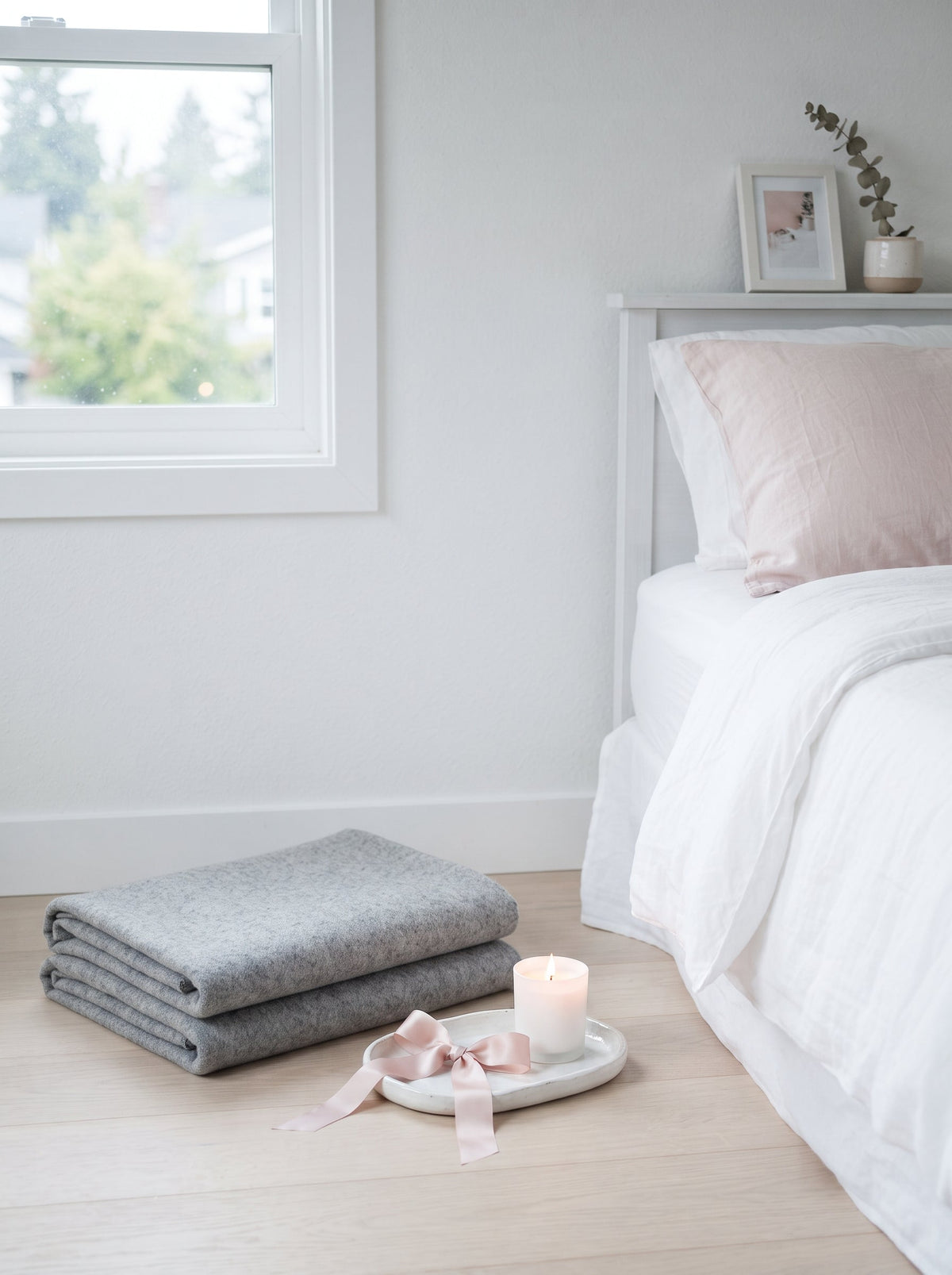 Folded grey blankets, a lit candle and a blush ribbon on a tray beside a white bed