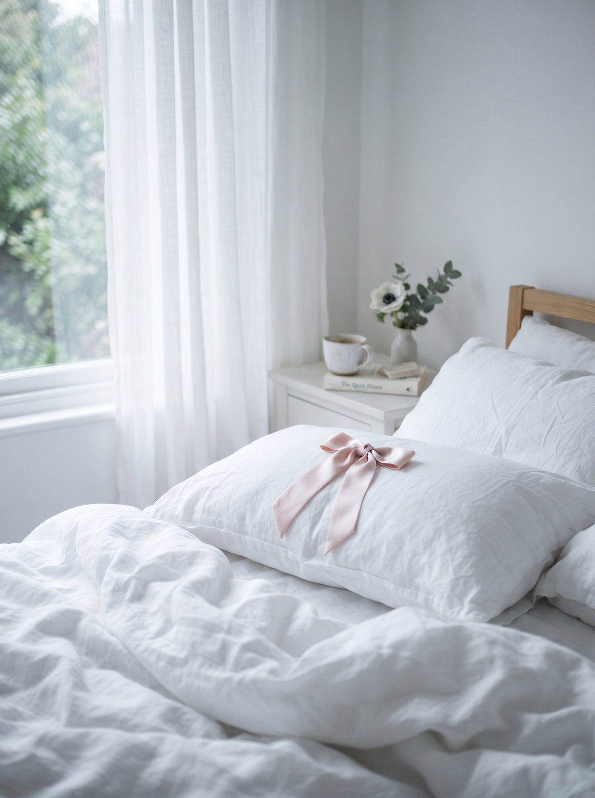 Morning light on white linen bedding with a blush satin ribbon tied on the pillow